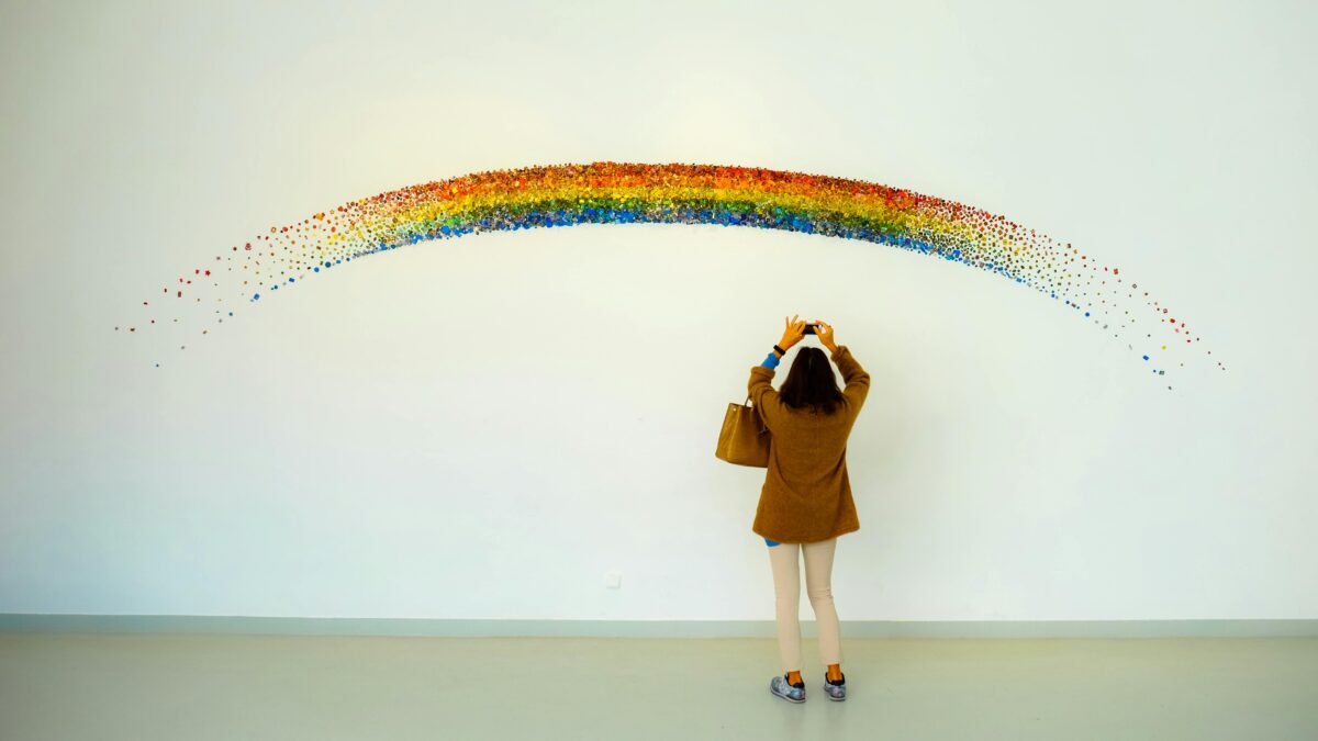 A woman stands observing a colorful rainbow art installation indoors.
