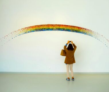A woman stands observing a colorful rainbow art installation indoors.